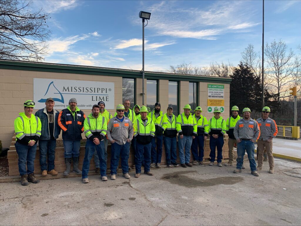 A team of employees in safety gear stand as a group in front of an office with a Mississippi Lime logo sign.